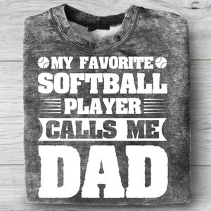 May include: A gray sweatshirt with the text "MY FAVORITE SOFTBALL PLAYER CALLS ME DAD" in white. The design includes softball graphics. The shirt is folded on a light wood surface.