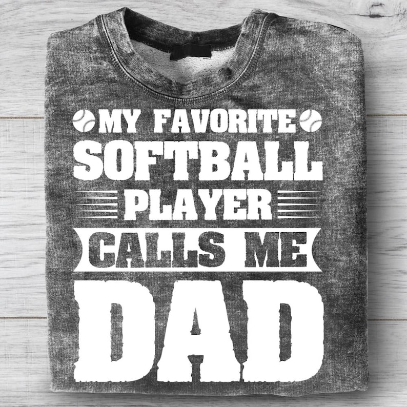 May include: A gray sweatshirt with the text "MY FAVORITE SOFTBALL PLAYER CALLS ME DAD" in white. The design includes softball graphics. The shirt is folded on a light wood surface.
