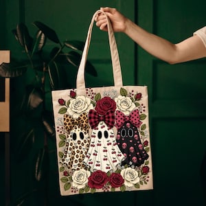 May include: A beige canvas tote bag featuring a graphic print of three ghosts wearing large bows. The ghosts are decorated with leopard print, cherry patterns, and pink skulls, surrounded by a border of red and white roses and green leaves.