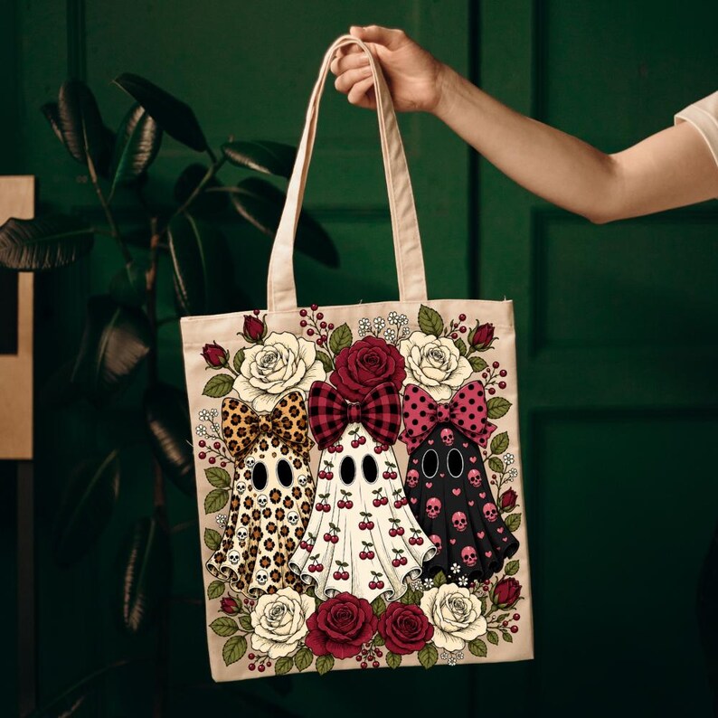May include: A beige canvas tote bag featuring a graphic print of three ghosts wearing large bows. The ghosts are decorated with leopard print, cherry patterns, and pink skulls, surrounded by a border of red and white roses and green leaves.