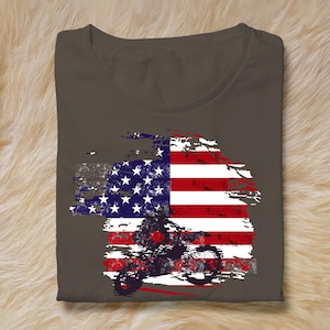 May include: A folded, dark gray t-shirt featuring a distressed American flag design. The flag graphic incorporates a silhouette of a motorcycle rider. The design is in shades of red, white, blue, and gray.