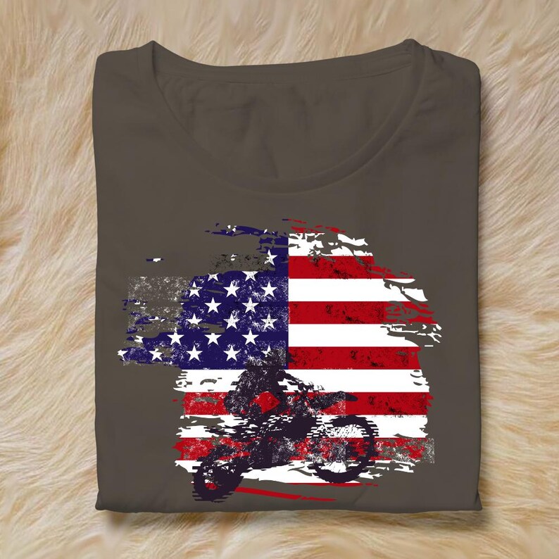 May include: A folded, dark gray t-shirt featuring a distressed American flag design. The flag graphic incorporates a silhouette of a motorcycle rider. The design is in shades of red, white, blue, and gray.