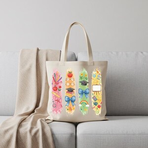 May include: A beige tote bag featuring four vertical watercolor designs with school-themed illustrations. The designs include pencils, apples, books, graduation caps, bows, rainbows, and geometric shapes. The bag is sitting on a gray couch.