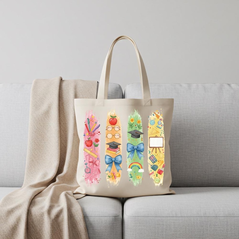 May include: A beige tote bag featuring four vertical watercolor designs with school-themed illustrations. The designs include pencils, apples, books, graduation caps, bows, rainbows, and geometric shapes. The bag is sitting on a gray couch.