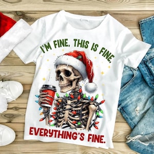 May include: White t-shirt featuring a skeleton wearing a Santa hat and wrapped in Christmas lights, holding a red coffee cup. The shirt has the phrases "I'm fine, this is fine" and "Everything's fine" printed on it.