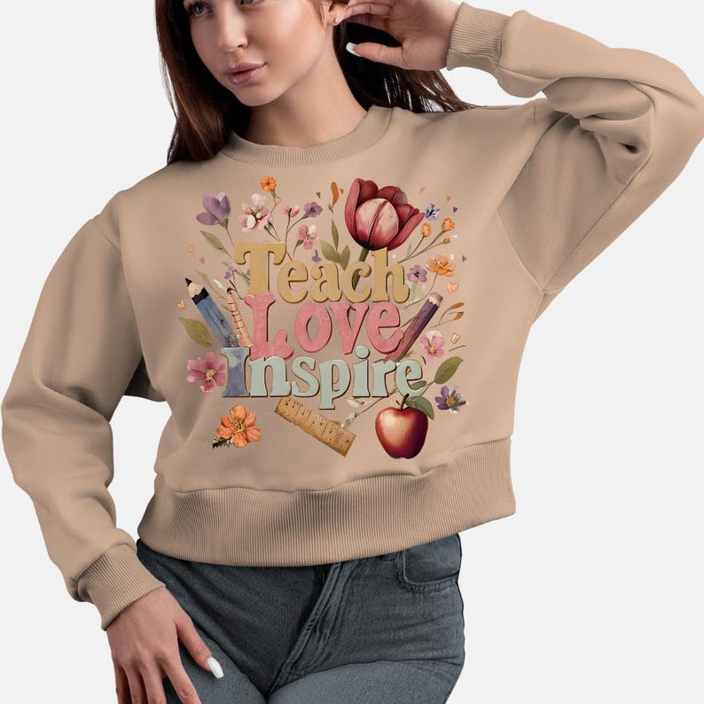 May include: Tan sweatshirt with the words "Teach Love Inspire" in colorful letters, surrounded by floral and school-themed graphics. Includes pencils, a ruler, and an apple. The sweatshirt has long sleeves and a crew neck.