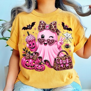 May include: A mustard yellow t-shirt featuring a graphic of a pink ghost wearing leopard print heart-shaped glasses and a matching bow. The ghost holds a pink drink and makes a peace sign, surrounded by bats, a spider web, pumpkins, and a handbag.
