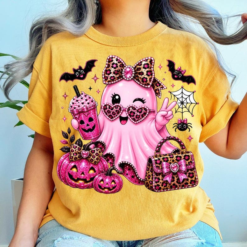 May include: A mustard yellow t-shirt featuring a graphic of a pink ghost wearing leopard print heart-shaped glasses and a matching bow. The ghost holds a pink drink and makes a peace sign, surrounded by bats, a spider web, pumpkins, and a handbag.