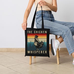 May include: Black tote bag with the text "THE CHICKEN WHISPERER" and a rooster illustration. The design features a retro color scheme with a rooster in the center, set against a backdrop of a forest and a sunset.