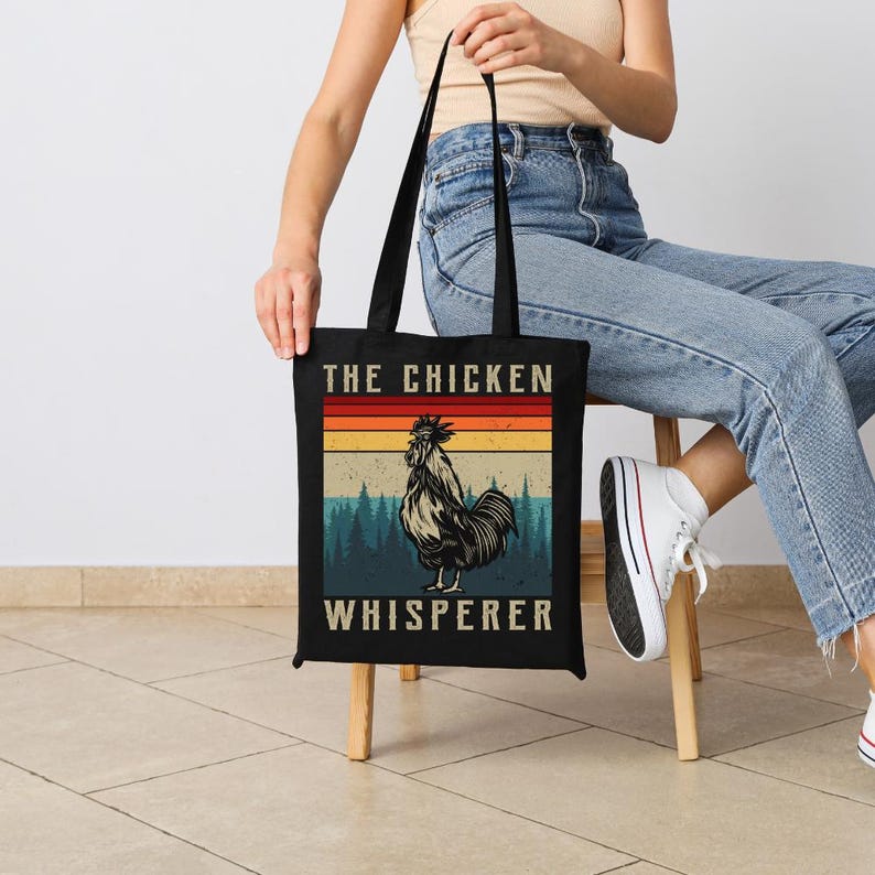 May include: Black tote bag with the text "THE CHICKEN WHISPERER" and a rooster illustration. The design features a retro color scheme with a rooster in the center, set against a backdrop of a forest and a sunset.