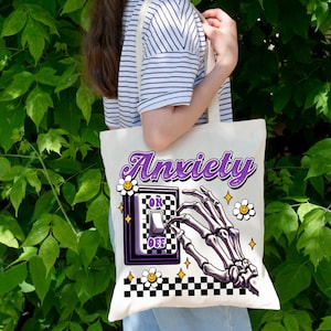 May include: Cream-colored tote bag with the word "Anxiety" in purple script. The design includes a skeleton hand reaching for a light switch with "OK" and "OFF" labels, surrounded by daisies and stars. A black and white checkered pattern borders the bottom.