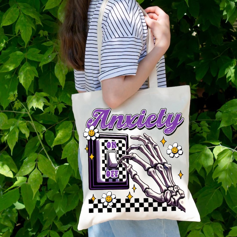 May include: Cream-colored tote bag with the word "Anxiety" in purple script. The design includes a skeleton hand reaching for a light switch with "OK" and "OFF" labels, surrounded by daisies and stars. A black and white checkered pattern borders the bottom.