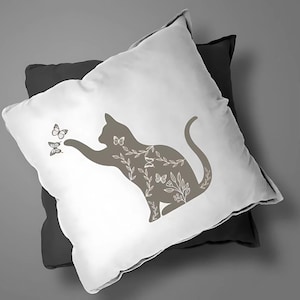 Pode incluir: Uma almofada branca com um design de silhueta de gato castanho. O gato est&aacute; rodeado de detalhes florais e borboletas. A almofada est&aacute; em cima de uma almofada cinzenta escura.