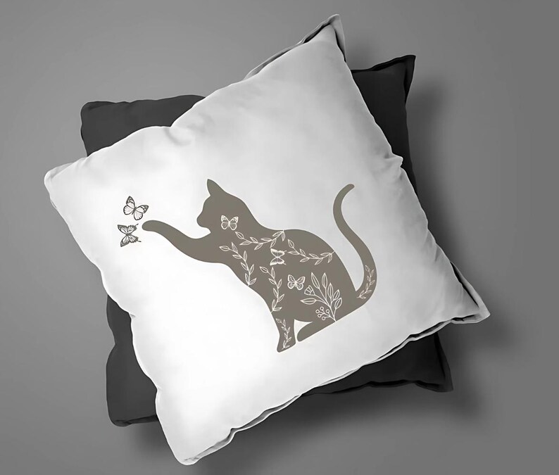 Pode incluir: Uma almofada branca com um design de silhueta de gato castanho. O gato est&aacute; rodeado de detalhes florais e borboletas. A almofada est&aacute; em cima de uma almofada cinzenta escura.