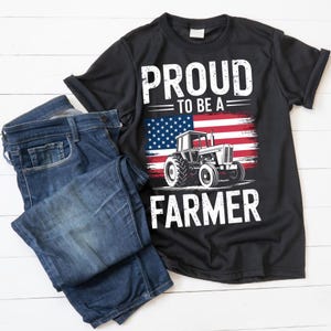 Puede incluir: Camiseta negra con el texto blanco "PROUD TO BE A FARMER". El dise&ntilde;o incluye una bandera estadounidense y un gr&aacute;fico de tractor. Un par de vaqueros azules est&aacute; doblado junto a la camiseta.