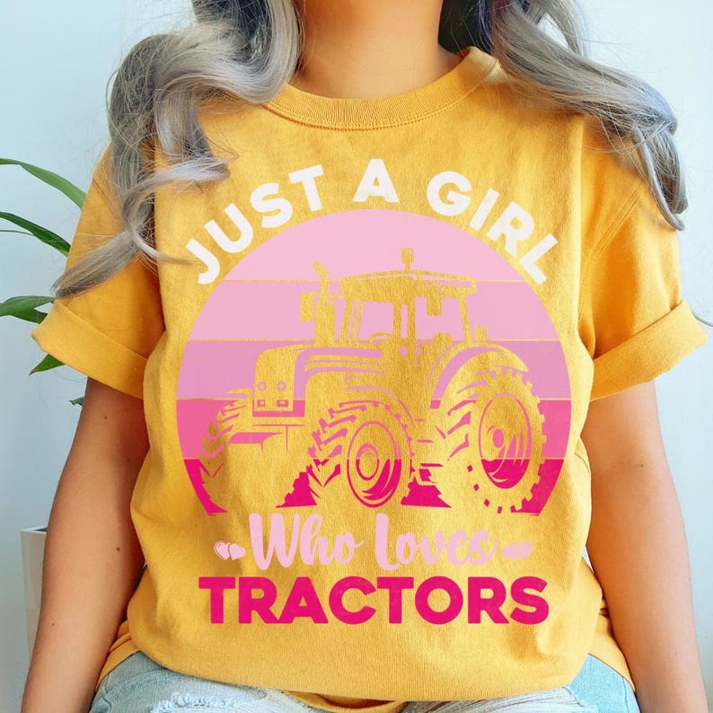 Puede incluir: Camiseta amarilla mostaza con un gr&aacute;fico de un tractor rosa dentro de un c&iacute;rculo rosa y blanco. El texto dice "JUST A GIRL Who Loves TRACTORS" en blanco y rosa.
