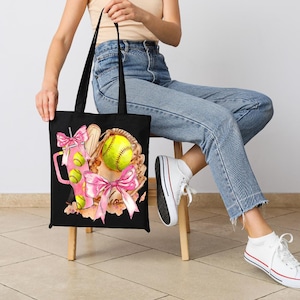 May include: Black tote bag featuring a softball theme. The design includes a softball, glove, and a pink drink with softball accents. Pink bows and a watercolor style add to the design. The bag has black handles.