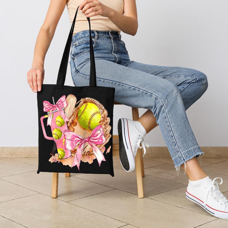 May include: Black tote bag featuring a softball theme. The design includes a softball, glove, and a pink drink with softball accents. Pink bows and a watercolor style add to the design. The bag has black handles.