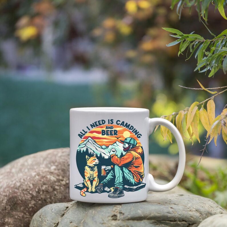 K&ouml;nnte beinhalten: Wei&szlig;e Keramik-Tasse mit Camping-Motiv. Die Tasse zeigt den Text "ALL I NEED IS CAMPING AND BEER" mit einer Illustration einer Person, einer Katze, Bergen und einem Sonnenuntergang. Die Tasse hat einen gebogenen Griff.