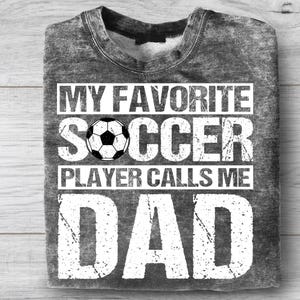May include: A gray sweatshirt with the text "MY FAVORITE SOCCER PLAYER CALLS ME DAD" in white. A soccer ball graphic is centered above the words "PLAYER CALLS ME". The sweatshirt is folded on a light wood surface.