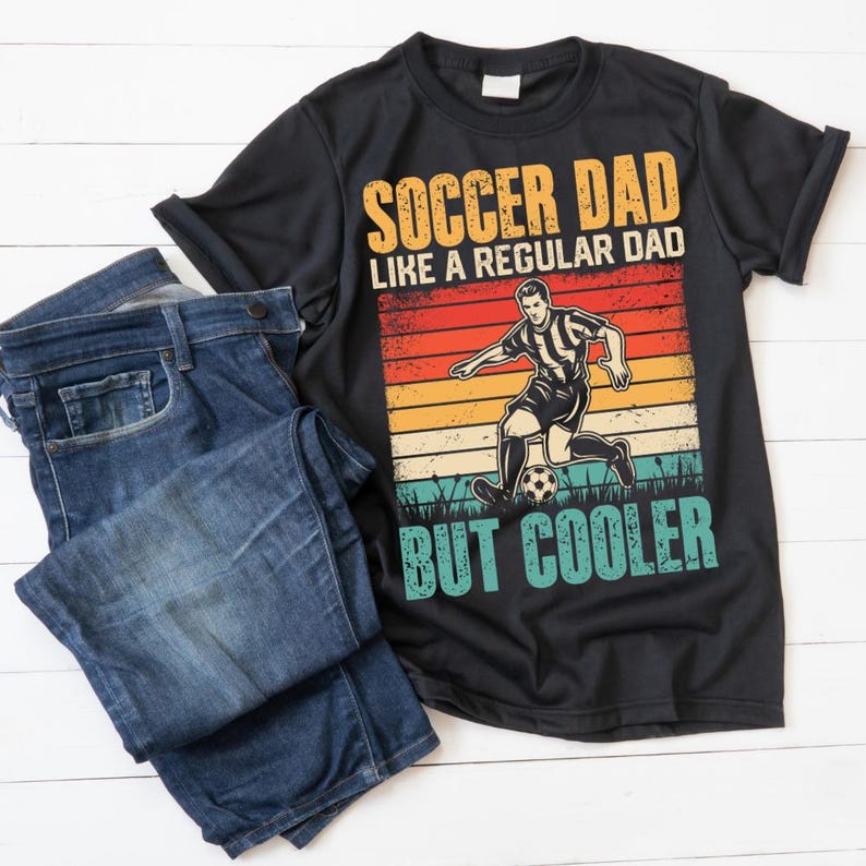 May include: Black t-shirt with the text "SOCCER DAD LIKE A REGULAR DAD BUT COOLER" in orange, yellow, and teal. The shirt features a soccer player graphic against a retro sunset design. Blue denim jeans are folded next to the shirt.