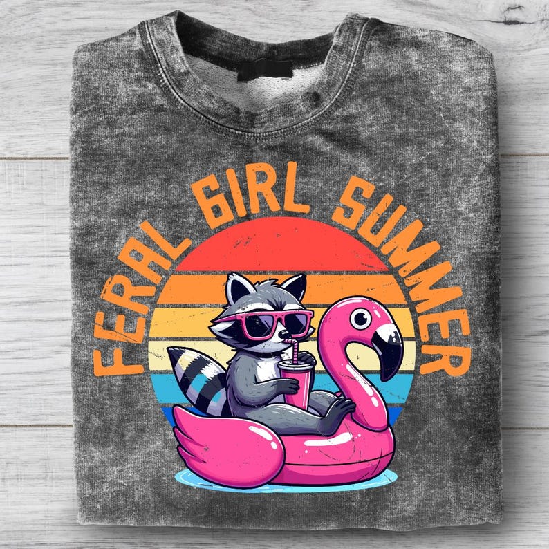 May include: A gray sweatshirt with a graphic of a raccoon wearing sunglasses and floating on a pink flamingo pool float. The text "FERAL GIRL SUMMER" is arched above the raccoon and flamingo, with a sunset background.