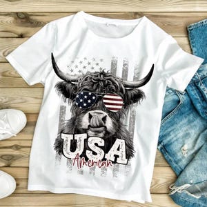 May include: White t-shirt featuring a black and white illustration of a Highland cow wearing sunglasses with the American flag design. The text "USA American" is printed below the cow image, with a distressed flag background.