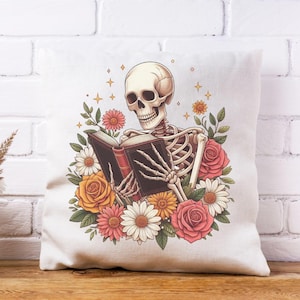 Puede incluir: Un coj&iacute;n cuadrado blanco con un dise&ntilde;o de un esqueleto leyendo un libro, rodeado de flores. El esqueleto se representa con un cr&aacute;neo y una caja tor&aacute;cica, sosteniendo un libro con lomo rojo. Las flores incluyen rosas y margaritas.