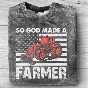 Puede incluir: Una sudadera gris con un aspecto desgastado, con un gr&aacute;fico de tractor rojo sobre una bandera estadounidense estilizada. El texto "SO GOD MADE A FARMER" est&aacute; impreso en blanco, con letras en negrita, creando un tema patri&oacute;tico y agr&iacute;cola.