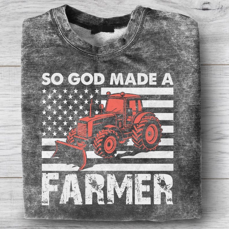 Puede incluir: Una sudadera gris con un aspecto desgastado, con un gr&aacute;fico de tractor rojo sobre una bandera estadounidense estilizada. El texto "SO GOD MADE A FARMER" est&aacute; impreso en blanco, con letras en negrita, creando un tema patri&oacute;tico y agr&iacute;cola.
