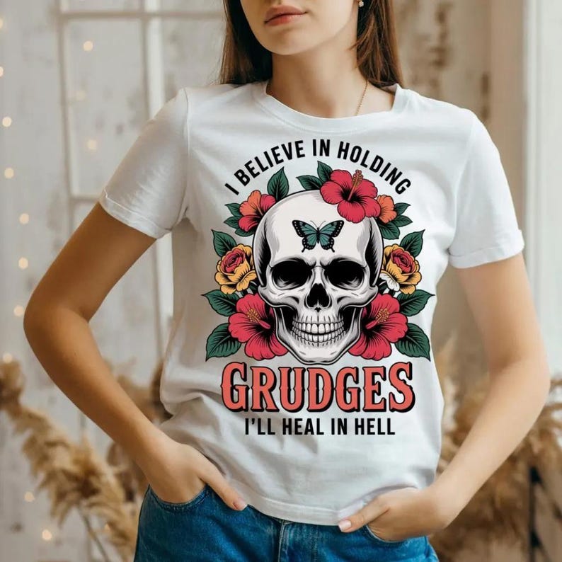 May include: White t-shirt featuring a skull design with a butterfly, surrounded by red hibiscus and yellow roses. The text reads "I BELIEVE IN HOLDING GRUDGES I'LL HEAL IN HELL".