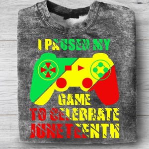 Pu&ograve; includere: Una felpa grigia con una grafica colorata di un controller per videogiochi in verde, giallo e rosso. Il testo "I PAUSE MY GAME TO CELEBRATE JUNETEENTH" &egrave; stampato sopra e sotto il controller in verde, giallo e rosso.