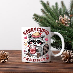 May include: White ceramic mug featuring a cartoon raccoon wearing a hat and holding a cup with whipped cream. The mug has the text "SORRY CUPID I'VE BEEN FERAL" in red. Pink hearts and arrows decorate the design. A fun gift for Valentine's Day.