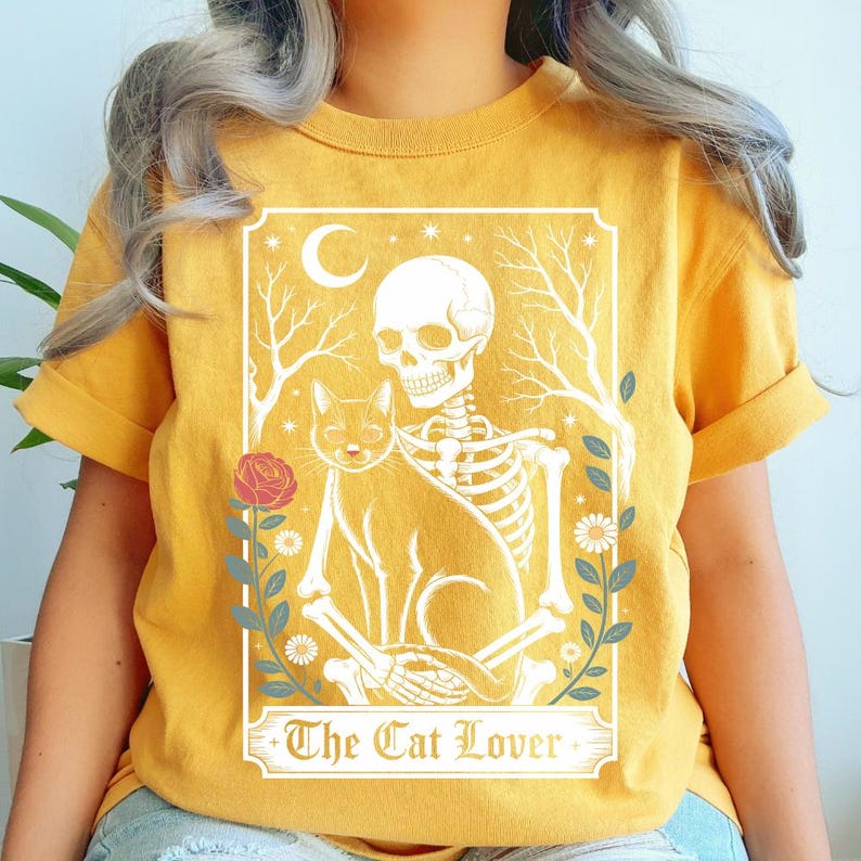 K&ouml;nnte beinhalten: Senfgelbes T-Shirt mit einer Grafik eines Skeletts, das eine Katze umarmt, mit einem Mond und floralen Akzenten. Der Text "The Cat Lover" ist unten auf dem Design zu sehen. Das T-Shirt hat hochgekrempelte &Auml;rmel.