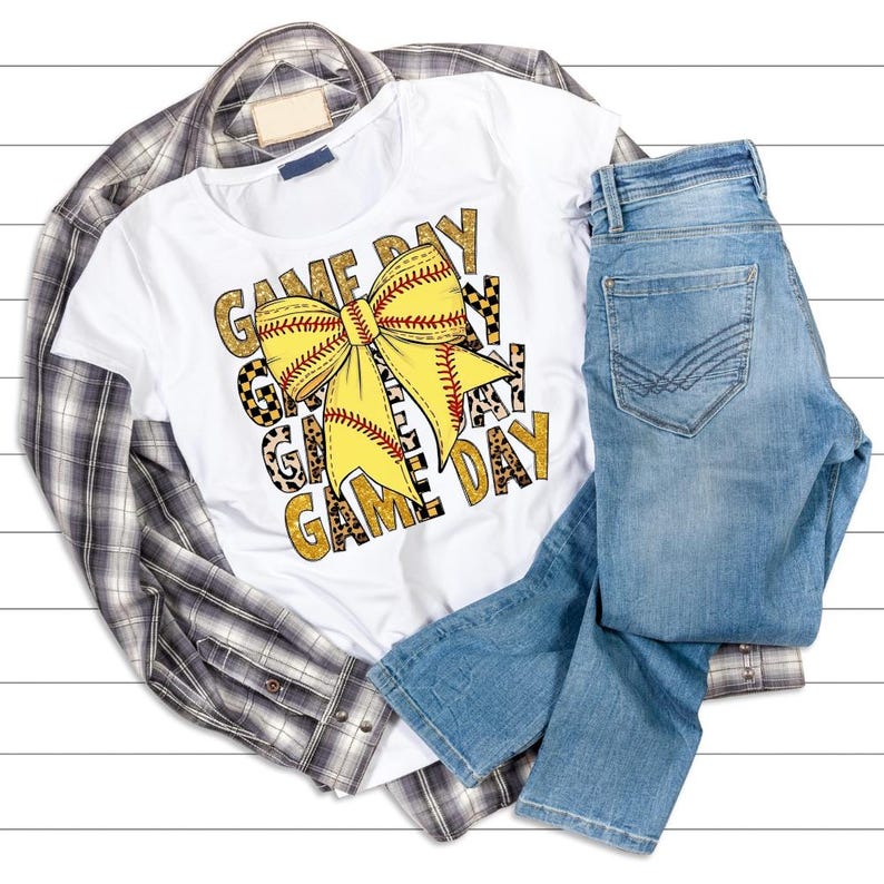 May include: A white t-shirt with a softball bow graphic and the words "GAME DAY" in gold and black. The shirt is paired with light wash denim jeans and a plaid button-down shirt.