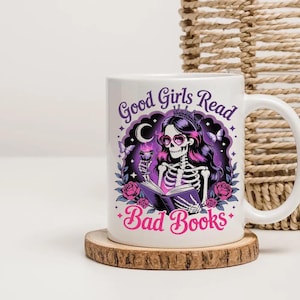 Puede incluir: Taza de cer&aacute;mica blanca con una ilustraci&oacute;n de un esqueleto con pelo rosa, gafas de coraz&oacute;n y una corona, sosteniendo una copa y leyendo un libro. La taza tiene el texto "Good Girls Read Bad Books" en morado y rosa.