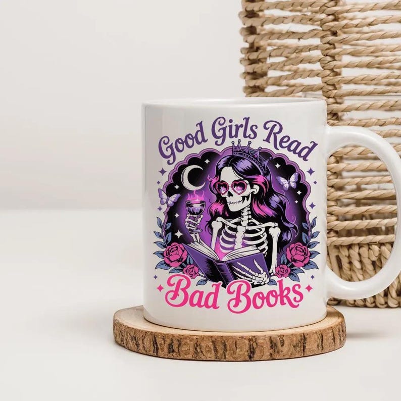 Puede incluir: Taza de cer&aacute;mica blanca con una ilustraci&oacute;n de un esqueleto con pelo rosa, gafas de coraz&oacute;n y una corona, sosteniendo una copa y leyendo un libro. La taza tiene el texto "Good Girls Read Bad Books" en morado y rosa.