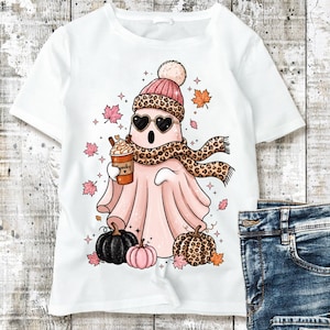 May include: A white t-shirt featuring a graphic of a light pink ghost wearing a leopard print scarf, a matching beanie, and heart-shaped sunglasses. The ghost holds a coffee cup labeled Spooky, surrounded by autumn leaves and three pumpkins.