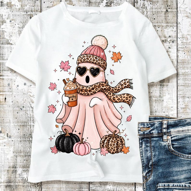 May include: A white t-shirt featuring a graphic of a light pink ghost wearing a leopard print scarf, a matching beanie, and heart-shaped sunglasses. The ghost holds a coffee cup labeled Spooky, surrounded by autumn leaves and three pumpkins.