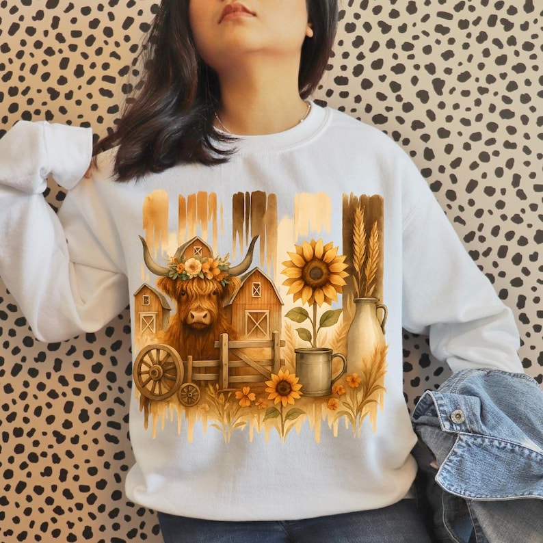 Puede incluir: Sudadera blanca con un dise&ntilde;o de acuarela de una vaca de las Tierras Altas con corona floral, graneros, girasoles y trigo. El dise&ntilde;o tiene un tema r&uacute;stico inspirado en la granja con tonos c&aacute;lidos y terrosos. El gr&aacute;fico est&aacute; centrado en la parte delantera.