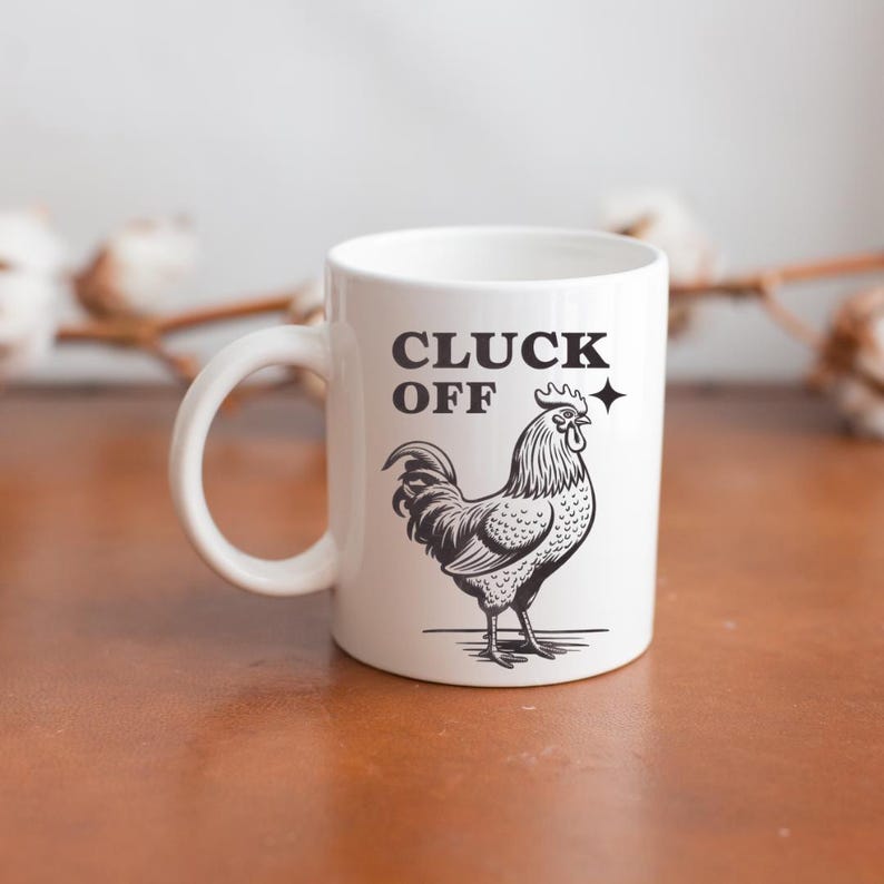May include: White ceramic mug with a black illustration of a rooster and the words "CLUCK OFF". The mug has a curved handle and is set against a blurred background.