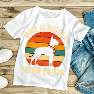 May include: White t-shirt with a graphic of a white dog wearing a scarf, set against a retro sunset design. The text "MINE IS BIGGER THAN YOURS" is printed above and below the dog illustration.