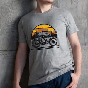 May include: A gray t-shirt featuring a vintage motorcycle graphic set against a sunset backdrop. The design includes a black motorcycle silhouette, orange and yellow sun, and mountain and tree outlines. The shirt is made of a soft material.
