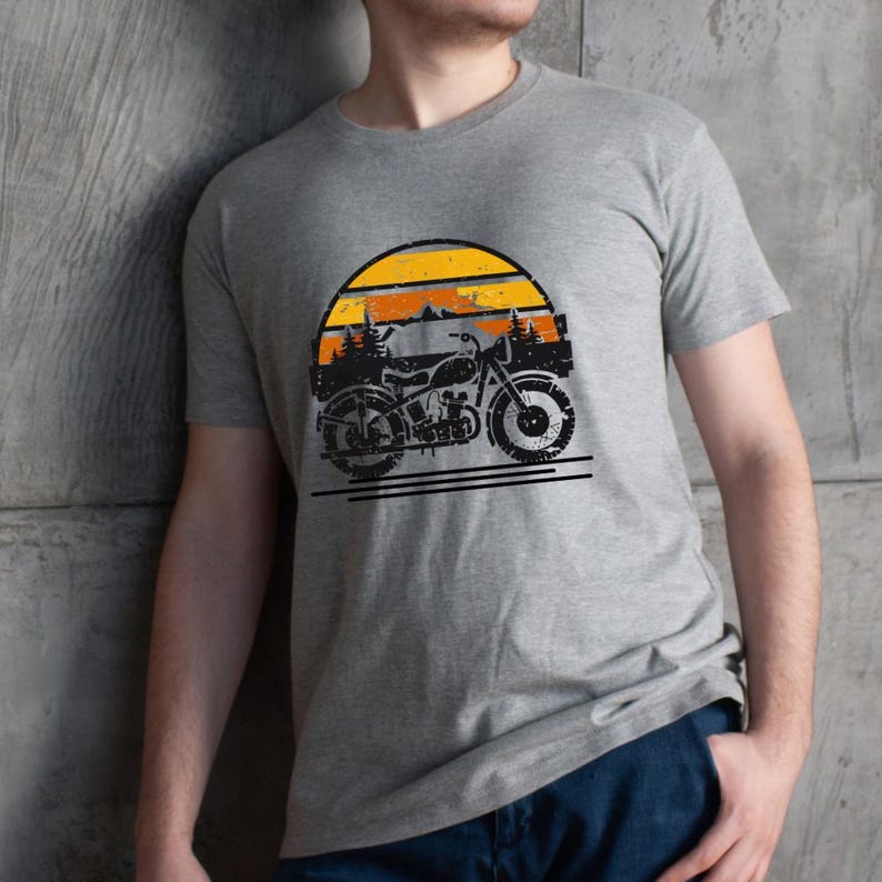 May include: A gray t-shirt featuring a vintage motorcycle graphic set against a sunset backdrop. The design includes a black motorcycle silhouette, orange and yellow sun, and mountain and tree outlines. The shirt is made of a soft material.
