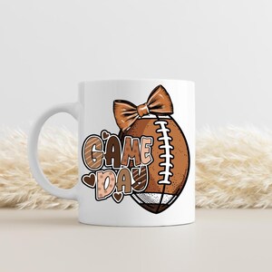 Puede incluir: Una taza de cer&aacute;mica blanca con un gr&aacute;fico de un bal&oacute;n de f&uacute;tbol americano marr&oacute;n con un lazo encima y el texto "GAME DAY" en letras marrones y beige. El dise&ntilde;o incluye peque&ntilde;os corazones y puntos marrones, con un asa en el lado izquierdo.
