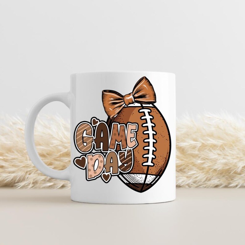 Puede incluir: Una taza de cer&aacute;mica blanca con un gr&aacute;fico de un bal&oacute;n de f&uacute;tbol americano marr&oacute;n con un lazo encima y el texto "GAME DAY" en letras marrones y beige. El dise&ntilde;o incluye peque&ntilde;os corazones y puntos marrones, con un asa en el lado izquierdo.