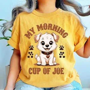 May include: Mustard yellow t-shirt featuring a cartoon puppy with the text "MY MORNING CUP OF JOE". The puppy has large eyes, a heart-shaped collar, and paw prints. The shirt has rolled-up sleeves.