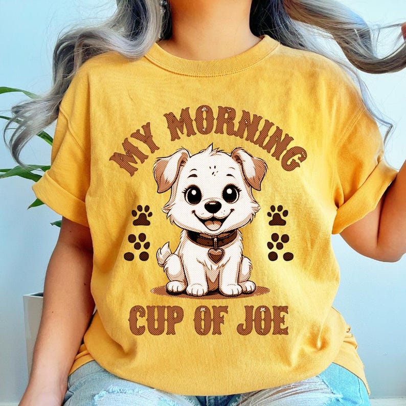 May include: Mustard yellow t-shirt featuring a cartoon puppy with the text "MY MORNING CUP OF JOE". The puppy has large eyes, a heart-shaped collar, and paw prints. The shirt has rolled-up sleeves.