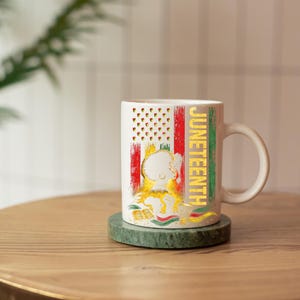 Pu&ograve; includere: Tazza in ceramica bianca con un design Juneteenth. La tazza presenta una bandiera americana stilizzata con strisce rosse e verdi, un'illustrazione dorata e la parola "Juneteenth" in lettere dorate. La tazza &egrave; appoggiata su un sottobicchiere di marmo verde.