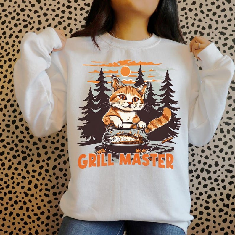 K&ouml;nnte beinhalten: Wei&szlig;es Sweatshirt mit einer Grafik einer rot-wei&szlig;en Katze, die Fisch grillt. Das Design beinhaltet einen Wald und einen Sonnenuntergang. Die Worte "GRILL MASTER" stehen in orangefarbenen Blockbuchstaben darunter.